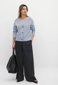 Cardigan azzurro chiaro con scollo a V, polsini a costine e tasche frontali, con bottoni dorati. Abbinato a pantaloni scuri a gamba larga e una borsa nera.