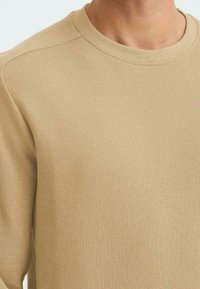 T-shirt beige a maniche lunghe in tessuto a coste testurizzato, con collo rotondo e dettagli di cucitura discreti lungo le cuciture delle spalle.