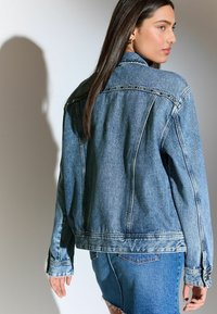 Chaqueta de mezclilla en azul claro con costuras laterales, detalles de botones y un corte ligeramente oversize. Presenta un cuello tipo punta y detalles de costura.