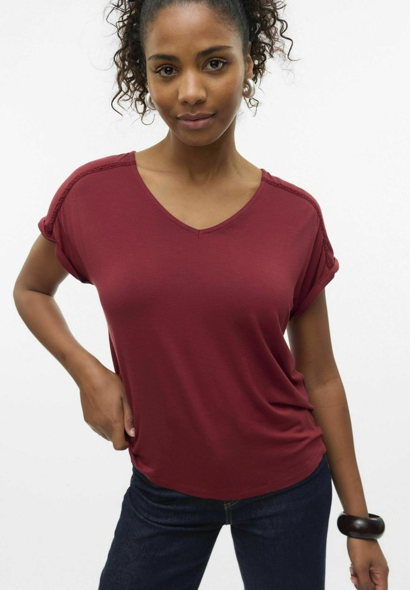 Camiseta de cuello en v burdeos con mangas cortas enrolladas, hecha de una tela suave. Llevada con jeans oscuros y una pulsera gruesa en la muñeca.