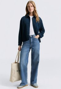 Dunkelblaues Cord-Hemd mit Knöpfen, weißes T-Shirt, hellblaue Jeans mit weitem Bein, beige Tragetasche mit Logo, hellblaue Schuhe.