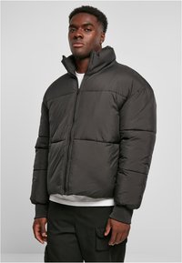 Chaqueta tipo plumón en negro, con un alto collar, cremallera frontal completa y silueta cuadrada con secciones cosidas para aislamiento.