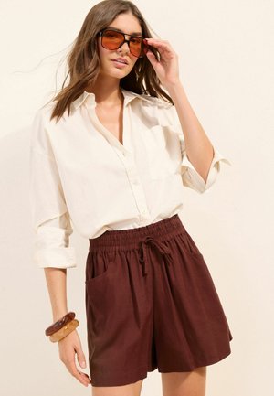 Woman wearing oversized cream button-up shirt, brown elastic-waist shorts with pockets, large amber sunglasses, and two chunky bracelets.