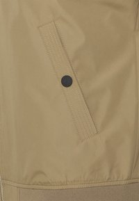 Chaqueta marrón con una tela suave y resistente al agua; presenta un bolsillo con solapa y un botón negro, además de detalles de costura visibles. Dobladillo elástico.