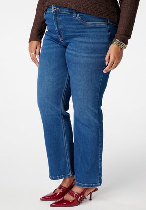 Blauwe spijkerbroeken met een rechte pijp, voorzakken en een subtiele vervaging. Gecombineerd met rode pumps met spitse neus en een bruine top met textuur.