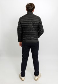 Giacca nera imbottita con collo alto, cuciture orizzontali e tessuto texturizzato. Indossata con pantaloni scuri e scarpe da ginnastica bianche.