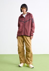 Sweatshirt burdeos com fecho e detalhes pretos e um bolso lateral, combinado com calças de carga em tom bege e sapatilhas beiges. Textura suave.