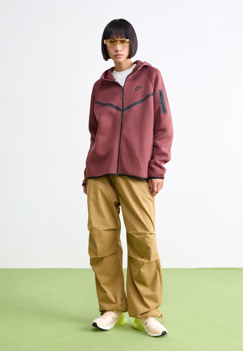 Sudadera con cremallera de color marrón burdeos con detalles en negro y un bolsillo lateral, combinada con pantalones cargo color beige y zapatillas beige. Textura suave.