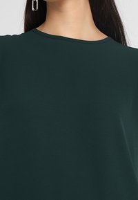 Top verde scuro con scollatura rotonda, texture liscia e maniche corte. Presenta un design minimale senza motivi o accenti prominenti.