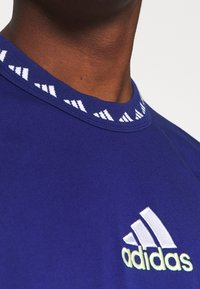 Camiseta atlética púrpura con cuello blanco acanalado que presenta acentos de logotipo repetidos. Mostrando un logotipo de Adidas bordado en blanco con detalles amarillos.