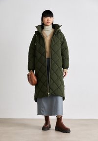 Groene gewatteerde lange jas met een rits, gecombineerd met een beige V-halstrui en een spijkerrok. Brown enkellaarzen en een bruine handtas maken de look compleet.