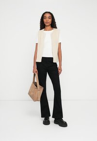 Beige ärmellose Weste über einem weißen T-Shirt, schwarze Schlaghosen und klobige schwarze Schuhe. Hält eine beige Handtasche; einfarbiger Hintergrund.