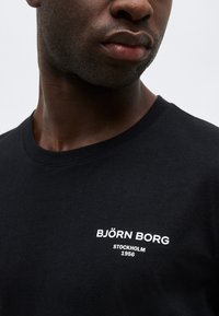 Κοντινό πλάνο ενός άνδρα που φοράει μαύρο T-shirt με λευκό κείμενο «BJÖRN BORG STOCKHOLM 1956» στο στήθος.