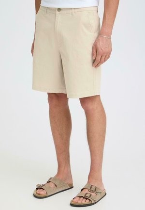 Męskie nogi w beżowych szortach do kolan i brązowych zamszowych sandałach z podwójnymi paskami na białym tle.