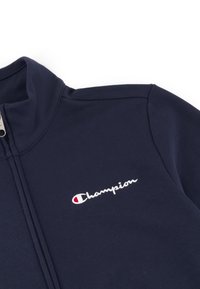 Sudadera navy de cremallera, hecha de tela lisa, con cuello alzado y logo bordado de "Champion" en blanco con acento rojo.