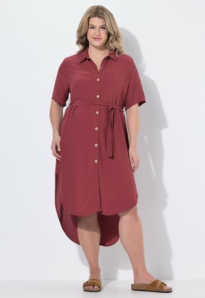 Kvinde iført en kortærmet, knappet, mørkerød kjole med taljesnor og brune slip-on sandaler, stående mod en hvid baggrund.