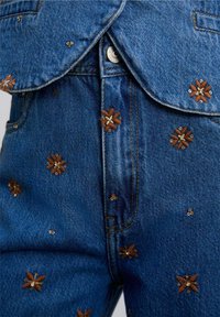 Farkut denim, jotka esittelevät monimutkaista ruskeaa kukka-embroiderya metallisilla yksityiskohdilla. Kangas on tummansininen, jossa on teksturoitu pinta ja näkyvät saumat.