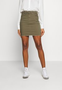 Falda de mezclilla verde oliva con silueta ajustada, botones frontales y dobladillo deshilachado. Combinada con zapatillas altas blancas.