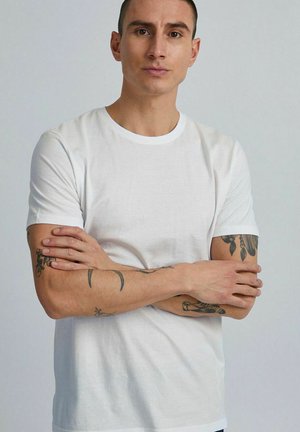 Mann mit kurzen Haaren und Tätowierungen auf beiden Armen, der ein einfaches weißes T-Shirt trägt und mit verschränkten Armen vor einem hellen Hintergrund steht.