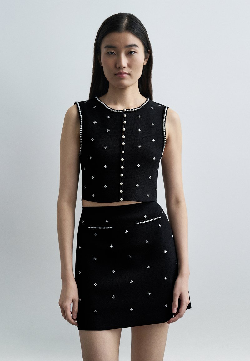 Γυναίκα με μακριά σκούρα μαλλιά που φοράει ένα μ αύρο αμάνικο crop top και φούστα που ταιριάζει, διακοσμημένα με μικρές χάντρες που μοιάζουν με πέρλες, τοποθετημένες σε σχέδια.