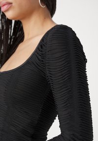Svart rynket bodysuit med scoop-hals og lange ermer, laget av teksturert materiale med horisontale rynker som skaper dybde.