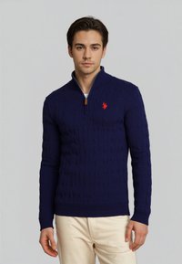 Mladý muž stojící v námořnickém modrém svetru s kulatým výstřihem a čtvrtinovým zipem, s červeným logem a krémovými kalhotami na jednoduchém pozadí.