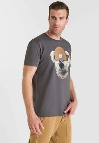 Camiseta de algodón gris con un gráfico de un oso que lleva un gorro marrón. Mangas cortas, cuello redondo estándar y corte relajado.