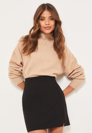 Femme aux cheveux bruns ondulés, portant un pull à col roulé beige et une mini-jupe noire taille haute avec une fente latérale, debout devant un fond uni.
