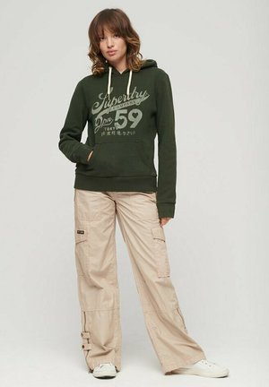 Olivgrüner Hoodie mit weißen Kordeln und bedrucktem Text, kombiniert mit beigen Cargo-Hosen mit Seitentaschen und verstellbaren Bündchen. Weiße Schuhe.
