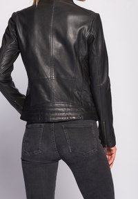 Žena v těsně přiléhajícím černém koženém sáčku se šitými detaily a tmavě šedých džínech, viděná zezadu na jednobarevném pozadí.