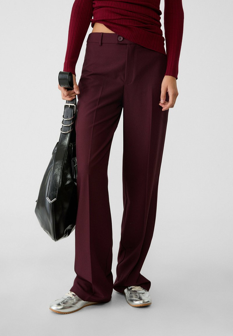 Pantaloni larghi bordeaux realizzati in tessuto morbido, con chiusura a bottoni e tasche laterali, abbinati a sneakers argento e a una borsa nera.