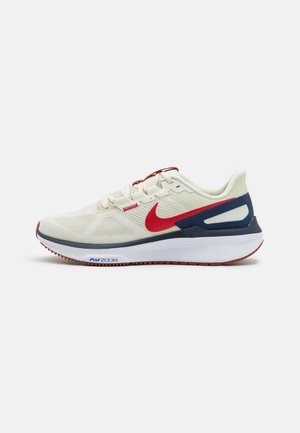 Chaussure de course beige clair avec swoosh Nike rouge, soutien du talon bleu marine, semelle intermédiaire blanche et semelle extérieure marron, vue de profil.