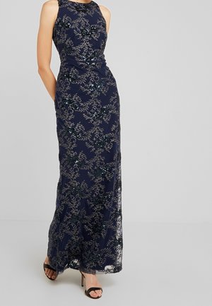 Robe longue sans manches bleu marine avec détails en dentelle florale et sequins, portée avec des sandales noires à talons ouverts.