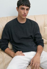 Junger Mann mit kurzen, dunklen Haaren, der einen dunkelgrauen Pullover und weiße Hosen trägt, sitzt auf einem beigen Sofa und schaut mit neutralem Gesichtsausdruck nach vorne.