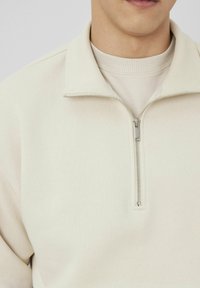 Sweatshirt zippé en beige clair, fabriqué à partir d'un tissu doux avec un col texturé. Il présente une fermeture éclair devant et des détails côtelés au niveau du col.