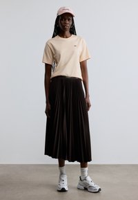 T-shirt en coton rose clair avec un petit logo vert, associé à une jupe plissée noire. Des baskets blanches et des chaussettes complètent la tenue.