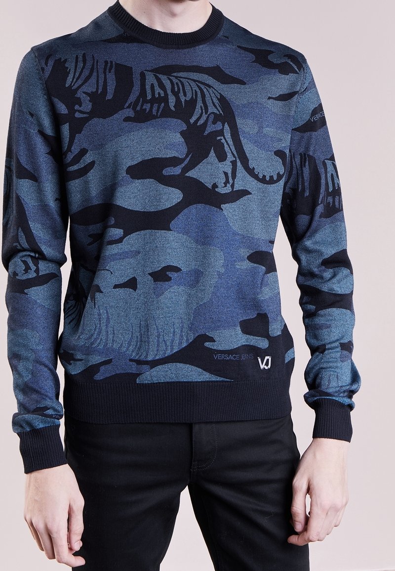 Pull en camouflage bleu présentant un motif de tigre, avec un col rond et des poignets côtelés, et un logo au bas. Fabriqué en tissu doux et texturé.