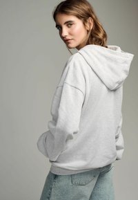 Hoodie grigio chiaro con una vestibilità rilassata, grande cappuccio e polsini a costine. Abbinato a jeans blu sbiaditi. Texture del tessuto morbida.