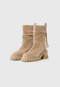 Bottes beige en daim mi-mollet avec talons blocs, détails cloutés sur les côtés, semelles en caoutchouc à crampons et accents en franges à l'arrière.