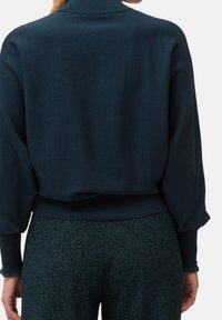 Suéter de punto verde oscuro con cuello alto, diseño oversized y dobladillo acanalado. Puños ajustados con detalle de borde deshilachado.