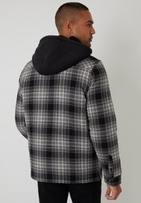 Schwarz-weiße karierte Jacke mit schwarzer Kapuze, die eine weiche Textur und geknöpfte Bündchen aufweist. Sicht von hinten, die das Design und die Schichtung zeigt.