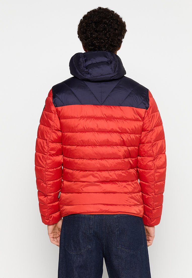 Chaqueta acolchada roja con capucha y hombros en azul marino. Presenta un acolchado horizontal, un corte relajado y costuras visibles.