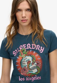 Donkerblauwe t-shirt met een grafisch ontwerp van een schedel omringd door een slang, omgeven door rozen en de tekst "SUPERDRY los angeles" in het roze.