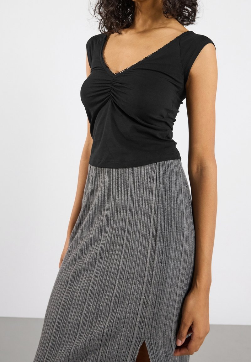 Femme portant un haut noir sans manches à encolure en V froncée et une jupe grise côtelée taille haute avec une fente à l'avant, debout devant un fond uni.
