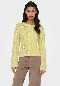 Cardigan tricoté jaune pâle avec un motif en dentelle texturée, boutons en forme de cœur et ceinture nouée à la taille. Manches longues avec poignets côtelés.