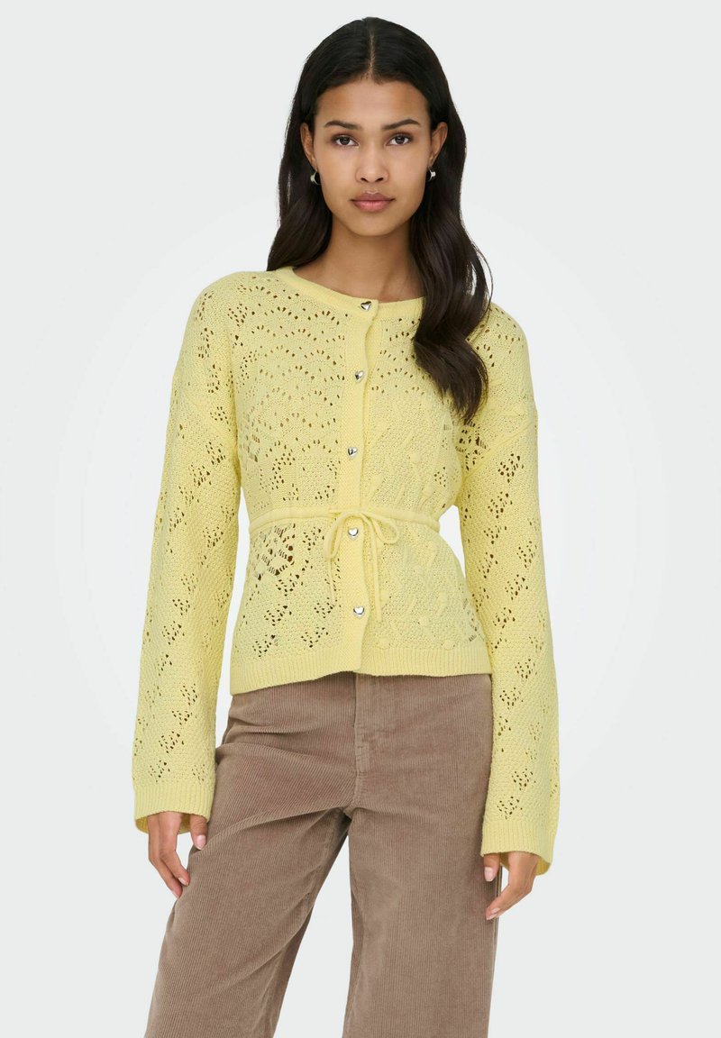 Cardigan tricoté jaune pâle avec un motif en dentelle texturée, boutons en forme de cœur et ceinture nouée à la taille. Manches longues avec poignets côtelés.