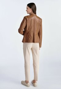 Brązowa skórzana kurtka o dopasowanym kroju i subtelnej teksturze, w połączeniu z jasnobeżowymi spodniami i neutralnymi sneakersami, widziana z tyłu.