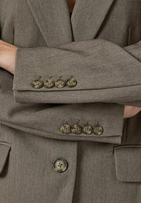 Bruine tweed blazer met een gestructureerde stof, vier decoratieve knopen op de manchetten en een enkele knoopsluiting aan de voorkant.