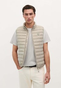 Lys beige polstret vest med høj krave, lynlås foran og lodrette syninger. Båret over en grå t-shirt med et lille logo på brystet.