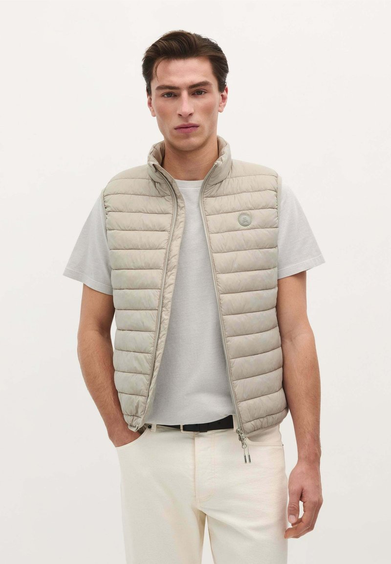 Lys beige polstret vest med høj krave, lynlås foran og lodrette syninger. Båret over en grå t-shirt med et lille logo på brystet.
